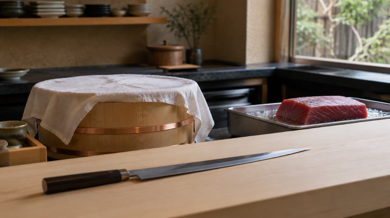 A pale hinoki counter with a long single-edged knife, a wooden rice tub under a damp cloth, and a slab of dark red tuna on a chilled tray.